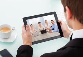 Motiv: Ein Mann schaut auf ein Tablet, auf dem andere Teilnehmer einer Konferenz beziehungsweise eines Webinars zu sehen sind. (Foto: apops - Fotolia.com)