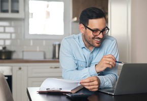 Motiv: Ein Mann sitzt an seinem Küchentisch und arbeitet lachend an einem Laptop. (Foto: shutterstock )
