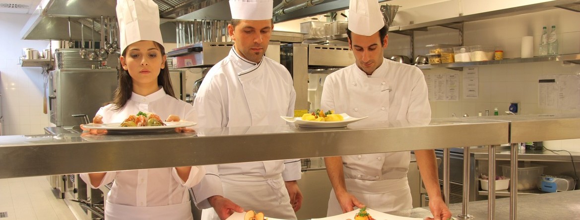Motiv: Zwei Köche und eine Köchin reichen in einer Küche fertig garnierte Teller zum Servieren an. (Foto: Fotolia )