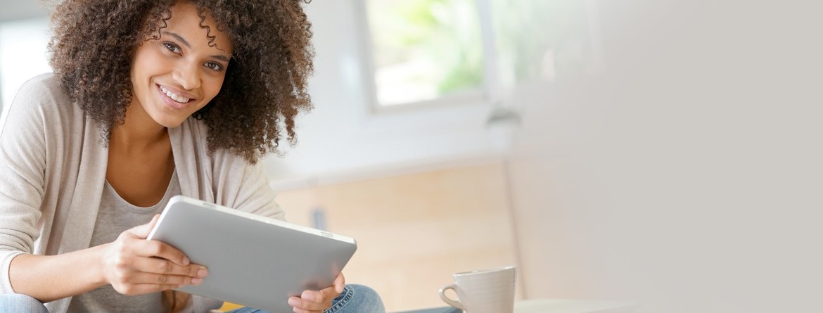 Motiv: Eine junge Frau sitzt lächelnd mit einem Tablet in der Hand und einer Tasse Kaffee vor sich in ihrem Zuhause. (Foto: Goodluz_AdobeStock)