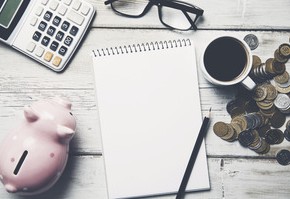 Motiv: Auf einem Tisch stehen ein Sparschwein und eine Kaffeetasse, liegen ein Notizblock mit Stift sowie mehrere Geldmünzen, eine Brille und ein Taschenrechner. (Foto: Tiko - Fotolia.com)