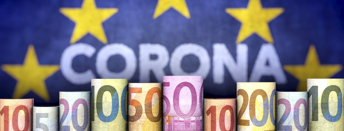 Motiv: Auf einer EU-Flagge steht im goldenen Sternenkreis das Wort "Corona". Davor sind zusammengerollte Euroscheine nebeneinander aufgestellt. (Foto: bluedesign - stock.adobe.com)