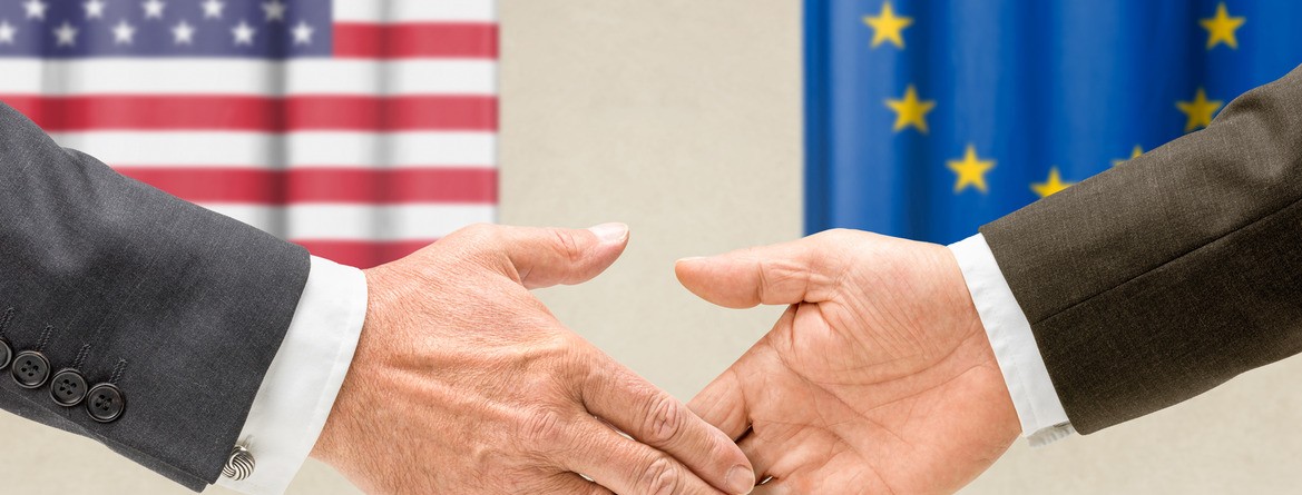 Motiv: Zwei Geschäftsmänner reichen sich vor den Flaggen der USA und der EU die Hände. (Foto: Zerbor - Fotolia.com)