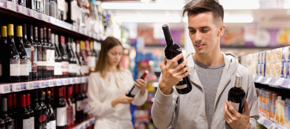 Motiv: junge Erwachsene betrachten Weinetikett einer Flasche vor einem Weinregal (Foto: JackF - stock.adobe.com)